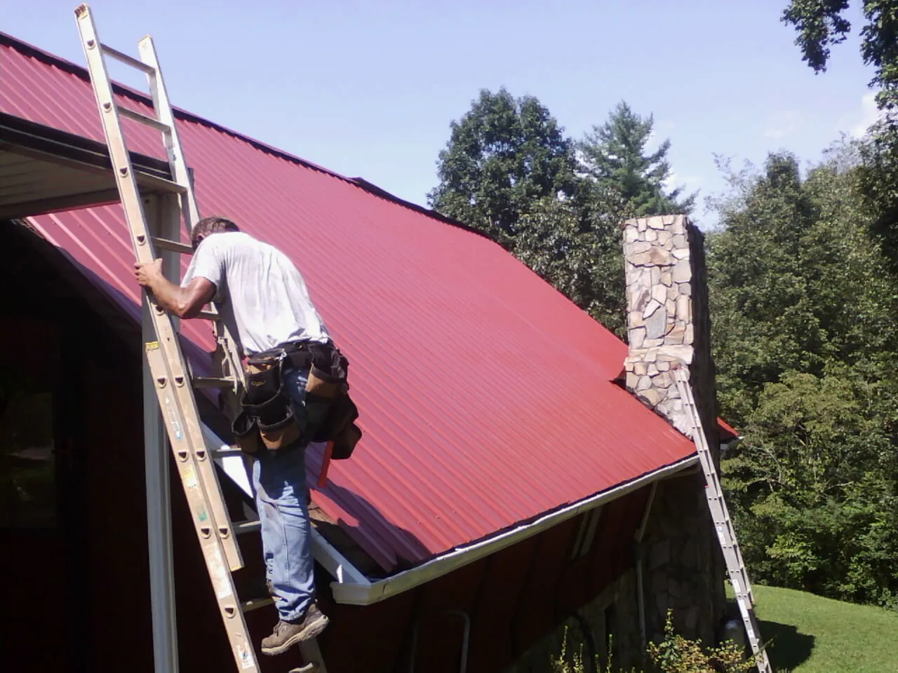 Roofer on a ladder beside a red metal roof and stone chimney — Soffit & Fascia Repair by Roofing Brothers in Milmay